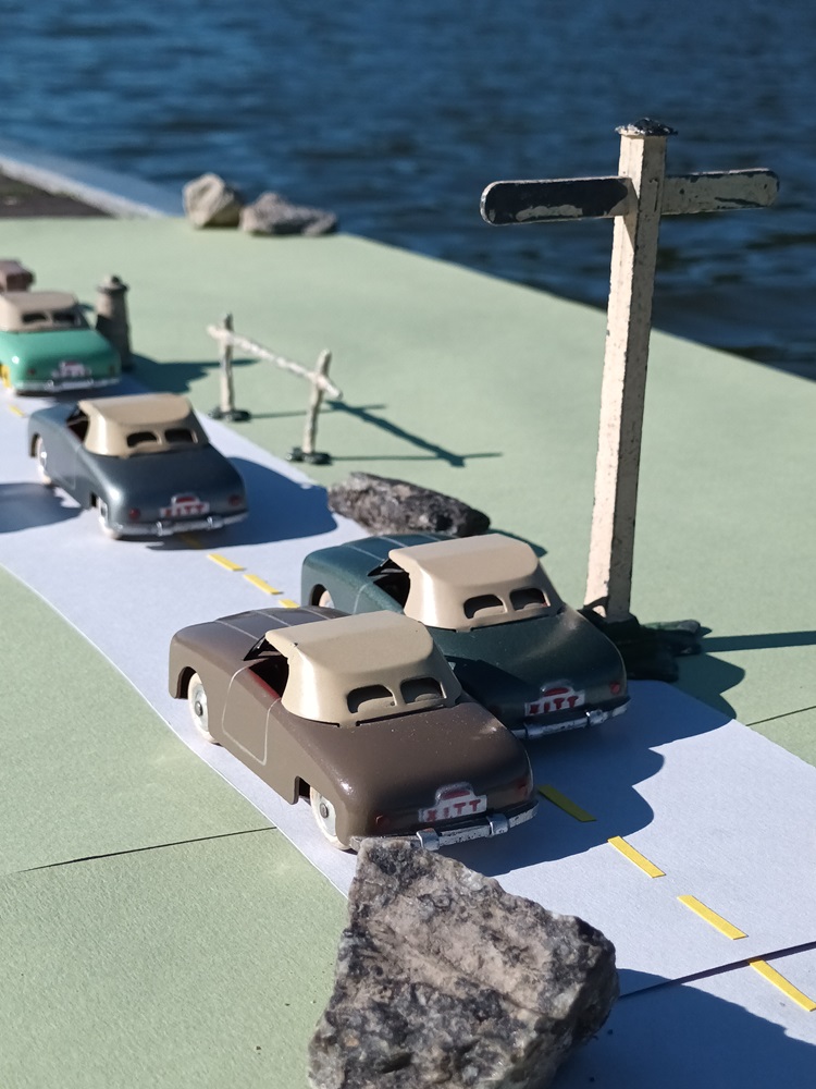 Vue plongeante de larrière de quatre Panhard Dyna Junior CIJ roulant dans une ligne droite sur une route au bord d'une rivière avec dépassement (3 modèles de premier type et une vert clair de second type)  